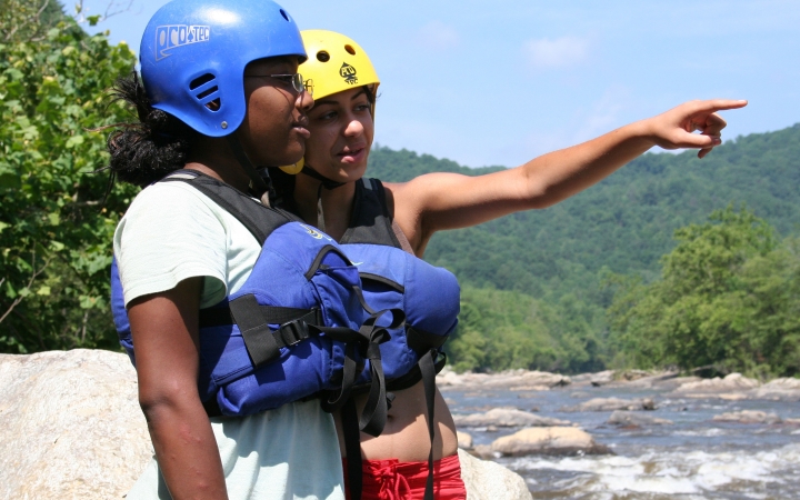 Two people wearing safety gear stand beside a river. One of them points out over the water.